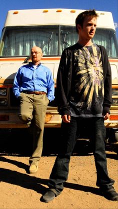 two men standing next to each other in front of a bus on dirt ground with blue sky and clouds behind them