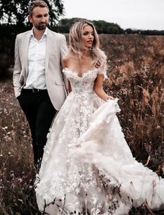 the bride and groom are walking through tall grass together in their wedding gowns that have flowers all over them