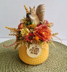 a squirrel figurine sitting on top of a yellow vase filled with flowers and leaves