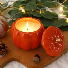 an orange pumpkin shaped candle sits on a cutting board next to some pine cones and acorns