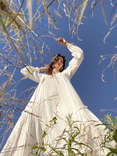 a woman in a white dress is standing among tall grass and trees with her arms outstretched