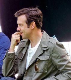 a man is sitting down talking on his cell phone while wearing an army green jacket