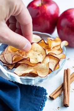 a hand picking up an apple chips from a bowl with cinnamon sticks and apples in the background