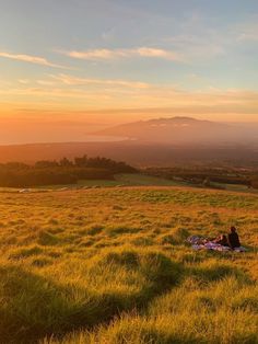 two people sitting on a blanket in the middle of a field at sunset with mountains in the distance