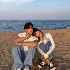 two people are sitting on the sand at the beach and one is holding his arm around the other