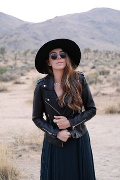 a woman wearing a black hat and dress in the desert with her hands on her hips