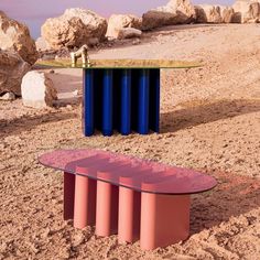 a pink bench sitting on top of a sandy beach next to rocks and a blue curtain