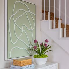 a white dresser topped with a vase filled with flowers next to a stair case and painting