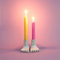 two candles with feet sticking out of them on a pink background, one is yellow and the other is pink