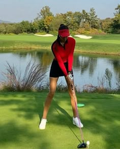 a woman in red shirt and black skirt playing golf on green field next to water