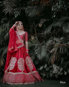 a woman in a red lehenga standing next to some palm trees and greenery