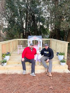 two men sitting on steps in front of a wooden deck with potted plants and flowers