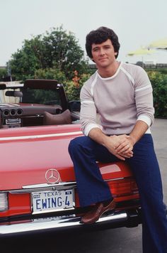 a man sitting on the hood of a red convertible car with his feet propped up