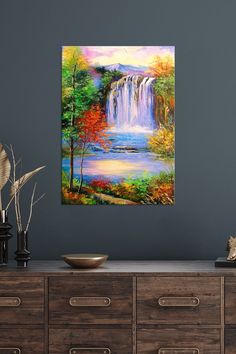 a painting of a waterfall with trees and water in the foreground, on a wall above a dresser