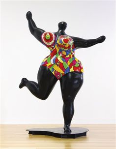 a statue of a woman in a colorful bodysuit is on a wooden table with a white wall behind it