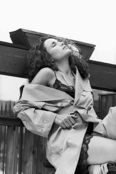 a woman sitting on top of a wooden bench wearing a trench coat and pearls in her hair