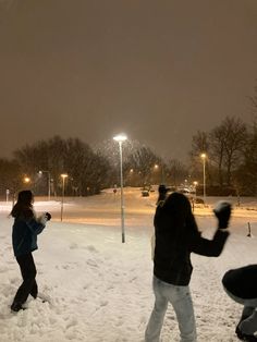 two people playing in the snow at night with street lights and cars on the other side