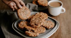 a person is picking up some cookies from a plate on a wooden table next to a cup of coffee