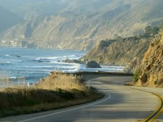 an empty road near the ocean with mountains on either side and water in the distance