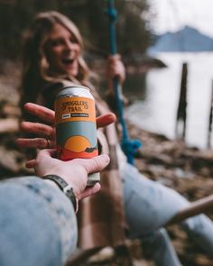 a woman holding up a can of beer in front of a lake with a rope