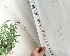 a hand is holding the edge of a curtain with beading on it and a potted plant next to it