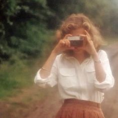 a woman taking a photo with a camera in front of her face and wearing a brown pleated skirt