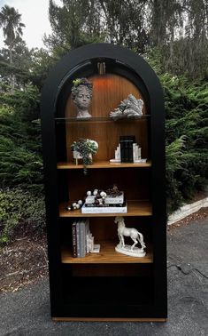 a book shelf with books and figurines on it in front of some bushes