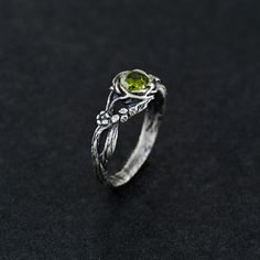 This sterling silver ring features a green gemstone cradled within a band of intertwining vines, reflecting the beauty of nature's organic forms. The gemstone symbolizes growth and renewal, perfect for those who love earthy, botanical designs. The oxidized finish highlights the rustic, vintage charm, making it an ideal statement piece or meaningful gift. Details:     Material: 925 Sterling Silver     Finish: Oxidized, highlighting its refined and timeless appeal     Stone: Peridot  Our pieces are crafted with love and meticulous attention to detail. We offer unique finish options that perfectly match your style:     Oxidized Finish: This gives the ring a noble, darkened patina reminiscent of vintage elegance, emphasizing its intricate details.     Shiny Finish: A mirror-like shine that pro Green Open Flower Ring For Promise, Nature-inspired May Birthstone Promise Ring, Nature-inspired Promise Ring With May Birthstone, Dark Green And Silver Ring, Nature-inspired Gemstone Flower Promise Ring, Green Gemstone Rings With Nature-inspired Style, Green Gemstone Rings In Nature-inspired Style, Nature-inspired Green Gemstone Rings, Green Sterling Silver Flower Ring