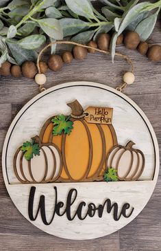 a welcome sign with pumpkins hanging from the side on a wooden table next to some leaves