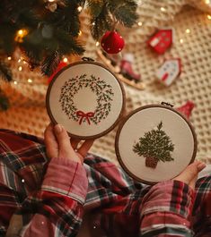 someone is holding up two cross stitch christmas ornaments in front of a christmas tree and stockings