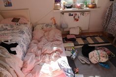 an unmade bed sitting next to a window in a room with lots of clutter on the floor