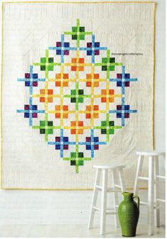 a white wall with a colorful quilt hanging on it's side and a green vase in the foreground