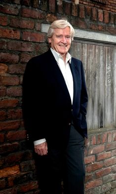 an older man standing in front of a brick wall wearing a black suit and white shirt