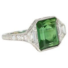 An Art Deco tourmaline, diamond, and platinum ring featuring an emerald-cut green tourmaline center flanked by trapezoid diamonds, with diamond-set shoulders and open scrolled foliate work in the gallery, in platinum. Marcus & Co. Atw tourmaline 4.61 cts., atw diamonds 1.75 cts. * Includes letter of authenticity for insurance purposes * Free shipping * Please do not hesitate to request additional photos of specific details or of this gorgeous period ring on a live model. Green Platinum Gemstones Fine Jewelry, Fine Green Platinum Gemstones Jewelry, Art Deco Green Ring, Gia Certified, Art Deco Green Ring Gia Certified, Art Deco Green Emerald Cut Diamond Ring, Green Emerald-cut Art Deco Diamond Ring, Fine Jewelry Green Diamond Platinum Ring, Gia Certified Green Emerald Art Deco Ring, Green Diamond Platinum Ring Fine Jewelry