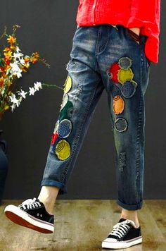 a woman in red shirt and jeans with flowers on her pants, standing next to a vase