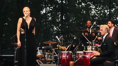 a woman in a black dress standing on stage with other people behind her and one man playing drums