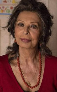 an older woman wearing a red sweater and necklace with beads on her neck, looking at the camera