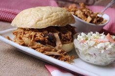 a pulled pork sandwich with cole slaw on a white plate next to a bowl of coleslaw