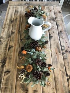 a wooden table topped with a white pitcher filled with pine cones and pumpkins on top of it