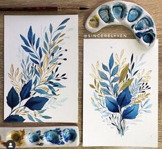 some blue and gold leaves on white paper next to watercolor paints, brush and acrylic