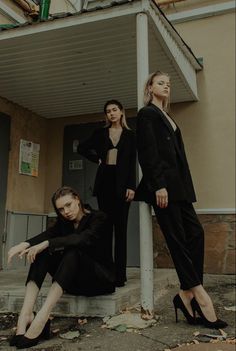 three women in black outfits sitting on the steps next to a building and one is leaning against a pole