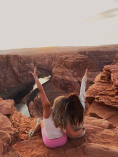 two women sitting on the edge of a cliff with their arms in the air