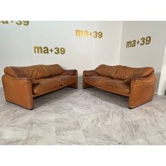 two brown leather couches sitting next to each other on a tile floor in front of a white wall