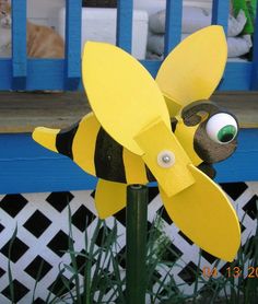 a yellow and black toy bee sitting on top of a green plant in front of a blue fence