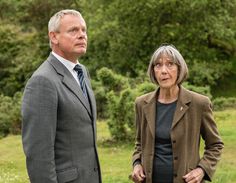 two people standing next to each other in front of green grass and trees with bushes behind them