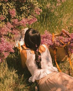 a woman sitting in the grass reading a book next to some flowers and a basket