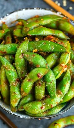 a white bowl filled with green beans next to chopsticks on a gray surface