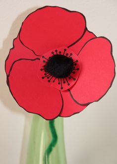 a red flower is in a green vase with white wall behind it and black center