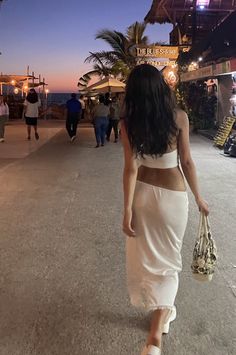 a woman in a white dress walking down the street at night with her back to the camera