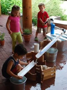 three children are playing in a water play area with buckets and barrels on the ground